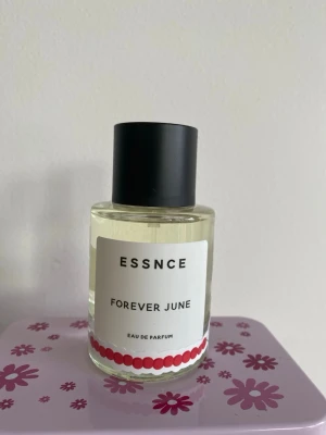ESSNCE Forever June parfym - Fräsch parfym från ESSNCE med namnet Forever June. Flaskan är rund och genomskinlig med svart lock och dekorerad med vita och röda etiketter samt jordgubbsdetaljer. Innehållet är ljust och flaskan har en modern, minimalistisk design. Testad 2 ggr. Dekorerad flaska 