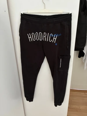 Svarta Hoodrich mjukisbyxor - Säljer ett par svarta mjukisbyxor från Hoodrich med vit och blå logga framtill. Byxorna har resår i midjan, muddar vid bensluten och en praktisk ficka på ena benet. Perfekta för en avslappnad och street-inspirerad stil.