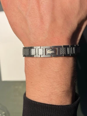 Silverfärgat armband från Lacoste - Snyggt armband i silverfärgat rostfritt stål från Lacoste med klassisk krokodil-logga. Länkarna har graverade detaljer som ger en cool och modern känsla. Perfekt accessoar för att lyfta din stil.