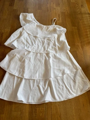 Vit oneshoulder klänning Gina Tricot - Superfin vit klänning från Gina Tricot med oneshoulder och volangdetaljer i flera lager. Perfekt för sommarens alla tillfällen. Lätt och luftig modell med smal axelrem och asymmetrisk skärning.