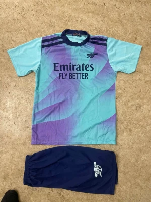 Arsenal träningsset Saka #7 blå/lila - Snyggt Arsenal träningsset med ljusblå och lila t-shirt samt mörkblå shorts. Tröjan har tryck med 'Emirates Fly Better' och Arsenal-logga, samt Saka och nummer 7 på ryggen. Perfekt för fotbollsträning eller match. Tillverkad i lätt och ventilerande material.