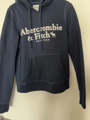 Mörkblå hoodie från Abercrombie & Fitch - Snygg mörkblå hoodie från Abercrombie & Fitch med vit broderad logga på bröstet och klassisk känguruficka. Tröjan har dragsko i huvan och är tillverkad i mjukt material som är skönt mot huden. Perfekt för en avslappnad och trendig look.