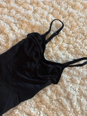 SKIMS - Svart body med axelband - Säljer en svart body från SKIMS XS med smala, justerbara axelband och lätt rynkad detalj vid bysten. Bodyn har en enkel och stilren design, perfekt att matcha med olika outfits. Materialet känns mjukt och följsamt. 