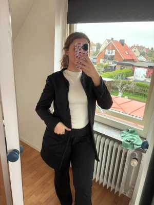 Svart lång kavaj med enkel design - Stilren svart kavaj med rak passform och långa ärmar. Kavajen har en minimalistisk look utan synliga knappar och går ner över höfterna. Perfekt för dig som gillar en clean och modern stil.