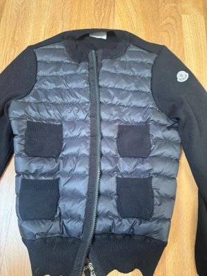 Svart cardiganjacka från Moncler - Snygg svart cardiganjacka från Moncler med quiltad framsida och stickade ärmar. Fyra stickade fickor framtill och dragkedja. Moncler-logga på ärmen. Perfekt för lager-på-lager och streetstyle. Pris kan diskuteras 