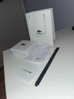 Svart armband från Lacoste - Snyggt svart armband från Lacoste med diskret och stilren design. Armbandet har en smal och rak form, tillverkat i ett tåligt material och kommer med originalförpackning, tygpåse och presentpåse. Perfekt accessoar för en modern look.