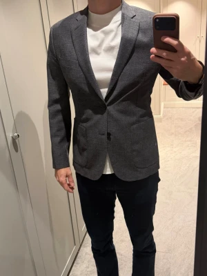 Grå kavaj från J.Lindeberg  - Säljer en stilren svart grå kavaj med slimmad passform och snygga detaljer som knappar på ärmsluten och två framfickor. Perfekt för dig som vill ha en snygg och tidlös look. Modell: Hopper 
