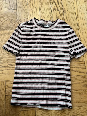 Randig ribbad t-shirt från H&M Divided - Säljer en ribbad t-shirt från H&M Divided i storlek S. T-shirten har rosa, svarta och vita ränder och är kortärmad med rund hals. Perfekt för en avslappnad och trendig stil.