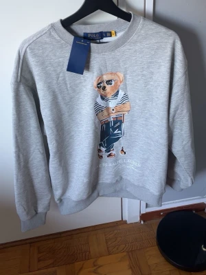 Grå sweatshirt Polo Ralph Lauren - Snygg grå sweatshirt från Polo Ralph Lauren med ikoniskt björntryck framtill. Storlek S men passar xs med. Pris kan diskuteras vid snabb affär.