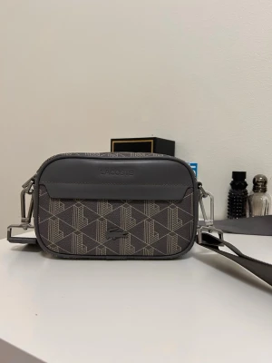 Grå axelväska från Lacoste - Snygg grå axelväska från Lacoste med geometriskt mönster och justerbar axelrem. Väskan har en framficka med Lacoste-logga och detaljer i silverfärgad metall. Perfekt för att bära dina viktigaste saker med stil.