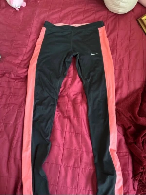 Nike leggings med rosa sidor - Snygga svarta leggings från Nike med breda rosa paneler längs sidorna. De har en tight passform och är perfekta för träning eller chill. Materialet är stretchigt och följsamt, med Nike-logga på låret. Supersköna och trendiga!