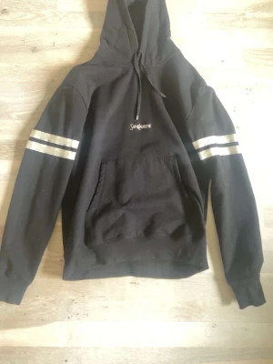 Svart hoodie från Saint Laurent - Svart hoodie från Saint Laurent med vit logga på bröstet och dubbla vita ränder på ärmarna. Klassisk känguruficka framtill och justerbar huva med snören. Mjuk bomullskänsla och avslappnad passform, perfekt för en cool streetstyle.