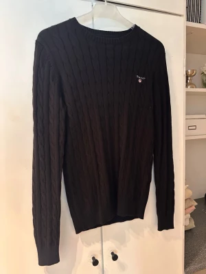 Svart kabelstickad tröja från Gant - Kabelstickad svart tröja från Gant med rund halsringning och diskret logga broderad på bröstet. Tröjan har långa ärmar och ribbade muddar vid ärmslut och nederkant. Perfekt för lager-på-lager och en klassisk stil.
