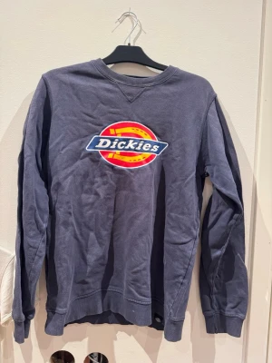 Blå Dickies sweatshirt med logga - Mörkblå sweatshirt från Dickies med stor broderad logga i rött, gult och vitt framtill. Tröjan har rund halsringning, långa ärmar och ribbade muddar vid ärmslut och nederkant. Perfekt för en avslappnad streetstil.