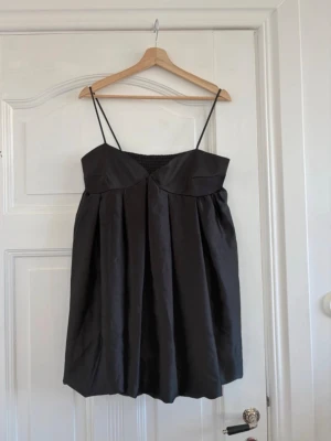 Svart kort klänning med tunna axelband - Svart kort klänning med tunna axelband och lätt puffig kjol. Klänningen har en enkel, stilren design med rynkade detaljer baktill och en något glansig finish. Perfekt för dig som gillar minimalistisk och trendig stil.