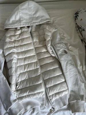 Vit pufferjacka med huva från Moncler - Snygg vit pufferjacka från Moncler med huva och dragkedja framtill. Jackan har quiltade paneler på framsidan och Moncler-logga på ärmen. Mjuka muddar och ficka med lock på ärmen. Perfekt för kalla dagar och riktigt stilren look. Loggan går och scanna , säljer för denne liten för mig jätte fin jacka och vänlig känd jackan behöver lite tvätt kan inte tvätta den för ska flytta 
