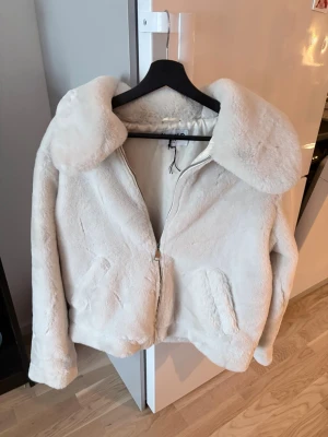 Vit teddyjacka från NA-KD - Säljer en fluffig vit teddyjacka från NA-KD med stor krage och dragkedja framtill. Jackan har två fickor på sidorna och är fodrad för extra värme. Perfekt för kalla dagar och ger en trendig look till din outfit.