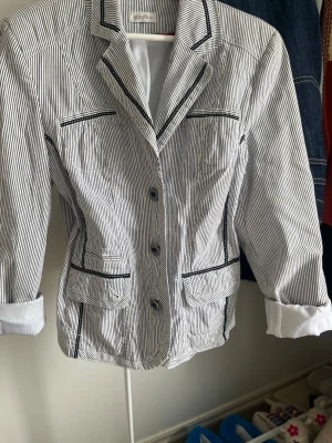 Randig kavaj  - Snygg kavaj med smala svarta och vita ränder från Bonita. Kavajen har klassisk krage, svarta kantdetaljer, knäppning framtill och fickor med lock. Ärmarna är uppvikta för en avslappnad look. Perfekt för att lyfta din outfit.