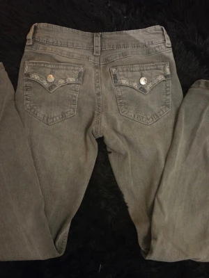 Grå jeans med broderade fickor - Snygga grå jeans med low rise, raka ben och två bakfickor med broderade detaljer och dekorativa silverfärgade knappar från Gina. Jeansen har en klassisk passform och är tillverkade i denim. Perfekta för dig som gillar unika detaljer på dina jeans.
