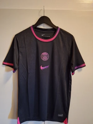 Svart PSG x Nike fotbollströja 2025 Special Edition  - Svart t-shirt från Nike i samarbete med Paris Saint-Germain. Tröjan har rosa och lila detaljer vid krage och ärmslut, samt PSG-logga och Nike Swoosh i rosa framtill. Tillverkad i ett lätt och sportigt material, perfekt för träning eller streetwear.