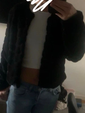 Svart pälsjacka i kort modell - Säljer en svart pälsjacka i kort modell med rund halsringning och diskret struktur. Jackan har en stilren look och passar perfekt till kalla dagar. Materialet är mjukt och ger en lyxig känsla.