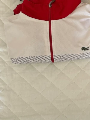 Röd och vit vindjacka från Lacoste - Snygg vindjacka från Lacoste i rött och vitt med dragkedja framtill. Jackan har ett diskret rutmönster längst ner och klassisk Lacoste-logga på bröstet. Perfekt för dig som gillar sportig stil.