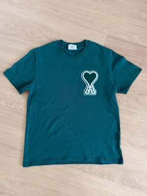 Grön t-shirt från Ami Paris med hjärtdetalj - Snygg grön t-shirt från Ami Paris med stor hjärtformad A-logga på bröstet. Klassisk rund hals och korta ärmar. Tillverkad i mjuk bomull som känns skön mot huden. Perfekt för en trendig och avslappnad look.