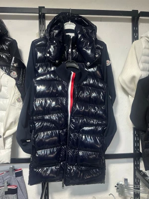Svart pufferjacka från Moncler - Snygg svart puffer kardigan från Moncler med glansig finish och stickade svarta ärmar. Jackan har dragkedja framtill, hög krage och Moncler-logga på ärmen. Röd och vit detalj vid dragkedjan ger en sportig touch. Perfekt för våren 