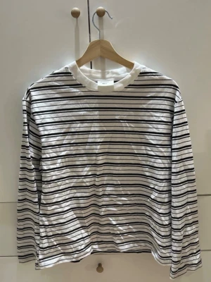 Gina tricot - Vit långärmad topp med svarta och mörkblå tunna ränder. Klassisk rak passform och rund halsringning. Tillverkad i mjuk bomull, perfekt för en avslappnad stil.