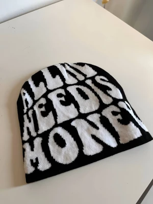 Svart y2k style mössa med vit text. - Säljer en svart mössa med stor vit text i grafisk stil där det står 'ALL I NEED IS MONEY'. Mössan är stickad och har en klassisk, rundad form. Perfekt accessoar för dig som vill sticka ut med ett statement-plagg. Mössan har dock ett utskriftsfel vid HE men annars är den i perfekt condition. 