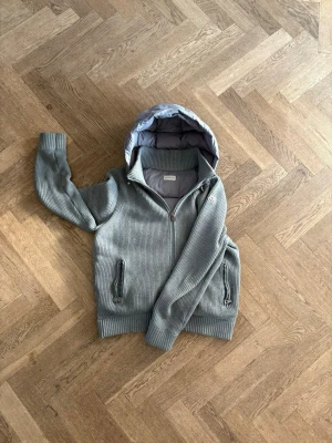 Grå Moncler Cardigan - Snygg grå stickad jacka från Moncler med quiltad huva och dragkedja framtill. Jackan har två fickor med dragkedja och ribbade muddar vid ärmslut och nederkant. Perfekt för dig som vill ha en stilren och bekväm look.