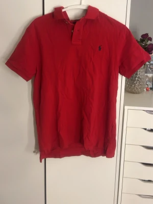 Röd pikétröja från Ralph Lauren - Röd pikétröja från Ralph Lauren med klassisk krage, korta ärmar och knappar vid halsen. Liten broderad logga på bröstet och tillverkad i mjuk bomull för en skön känsla.