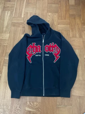 Svart zip hoodie från FSBN - Svart hoodie med dragkedja från FSBN. Stor röd och vit brodyrtext på bröstet med 'ESTD. 1992'. Mjuk fleece på insidan och klassisk känguruficka framtill. 