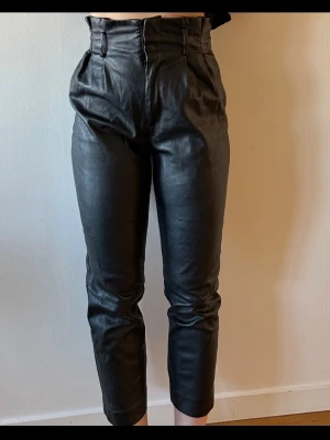 Svarta högmidjade croppade skinnbyxor - Stilrena svarta byxor i skinnimitation med hög midja och croppad längd. Byxorna har raka ben, bälteshällor och veck framtill för en modern och edgy vibe. Perfekta för dig som vill ha en trendig look.