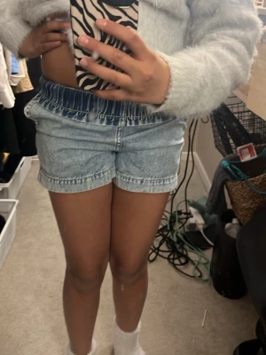 Ljusblå denimshorts med resårmidja - Säljer ett par ljusblå denimshorts med resår i midjan. De har en avslappnad passform och klassisk jeanslook, perfekta för varma dagar. Shortsen är utan synliga loggor och har en enkel, stilren design.