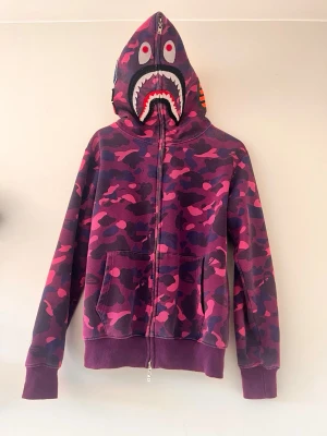 BAPE Shark Hoodie rosa camo - Säljer en ikonisk BAPE Shark Hoodie i rosa och lila camo med dragkedja hela vägen upp till huvan. Huvan har det klassiska hajansiktet och broderade bokstäver. Perfekt statement-plagg för streetwear-entusiasten.