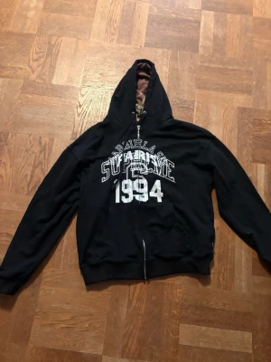 Svart Supreme x Margiela hoodie - Svart hoodie med dragkedja från Supreme och Maison Margiela. Stor vit print framtill med texten 'SUPREME', 'MARGIELA', 'PARIS' och '1994'. Klassisk huva med dragsko och känguruficka. Perfekt för dig som gillar streetwear och unika samarbeten.