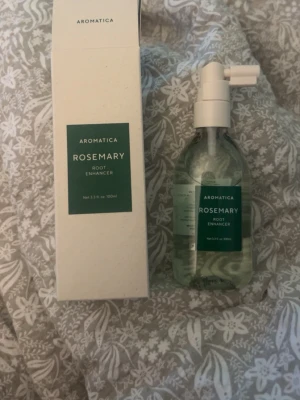 Aromatica Rosemary Root Enhancer - Aromatica Rosemary Root Enhancer är en hårvårdsprodukt i sprayform som kommer i en genomskinlig flaska med vit spraypump och grön etikett. Produkten innehåller rosmarin och är framtagen för att stärka hårbotten och rötterna. Förpackningen är stilren och modern. Helt oanvänd (nypris 259)