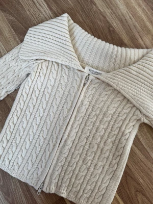 Beige kofta från Stradivarius - Kabelstickad beige kofta från Stradivarius med dragkedja framtill och bred ribbad krage. Koftan har långa ärmar och är tillverkad i ett mjukt stickat material som ger en trendig och mysig look. Jättesnygg för bara vardagen eller för att klä upp sig lite extra! Aldrig använd och priset kan diskuteras!!