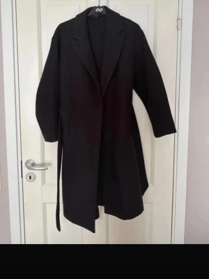 Svart lång kappa med skärp - Stilren svart lång kappa med klassisk krage och knytband i midjan. Kappan har raka ärmar och praktiska fickor på sidorna. Perfekt för dig som vill ha en enkel men snygg look till hösten eller vintern. katt i hemmet. nytvättad, rolla kappen behövs vill bara få sålt pris kan diskuteras 