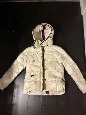 Vit dunjacka med huva från Moncler - Snygg vit dunjacka från Moncler med huva och coola detaljer som svarta knappar och dragkedjor. Jackan har Moncler-logga på bröstfickan och rödblå rand på insidan av huvan. Perfekt för kalla vinterdagar.