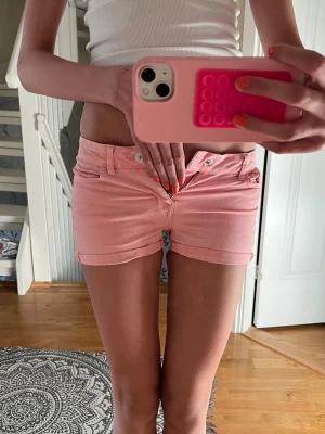 Lågmidjade rosa jeansshorts - Säljer ett par lågmidjade rosa jeansshorts med klassisk femficksdesign och uppvikta benslut. Shortsen har bälteshällor och stängs med knapp och dragkedja. Perfekta för varma dagar och en chill look. Dem passar xxs men dem är 134 men stora i storleken