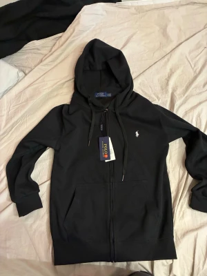 Svart hoodie från Polo Ralph Lauren - Svart hoodie från Polo Ralph Lauren med dragkedja framtill och klassisk huva. Helt ny. Tröjan har två fickor, ribbade muddar och den ikoniska vita logotypen broderad på bröstet. Perfekt för en avslappnad och stilren look.
