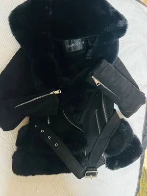Svart skinnjacka med fuskpäls från Garoff - Snygg svart skinnjacka från Garoff med stora fuskpälsdetaljer på krage och nederkant. Jackan har silvriga dragkedjor, bälte i midjan och en cool bikerstil. Perfekt för dig som vill sticka ut med en trendig look.