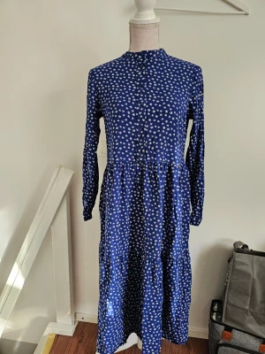 Blå blommig långklänning - En blå långklänning med små vita blommor, långärmad och med knappar framtill. Klänningen har en avslappnad passform och faller snyggt ner mot anklarna. Perfekt för dig som gillar en feminin och bekväm stil.