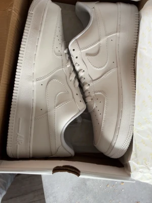 Nike Air Force 1 vita låga sneakers - Klassiska Nike Air Force 1 sneakers i helvitt läder. Modellen har en låg siluett, perforerad tåbox och den ikoniska swoosh-loggan på sidan. Tjock sula med AIR-dämpning för extra komfort. Perfekta för dig som gillar stilrena och tidlösa sneakers.