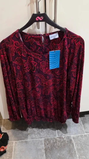 Mörkröd blommig blus från Zhenzi - Snygg mörkröd blus från Zhenzi med blommigt mönster i rött och svart. Blusen har rund halsringning och långa ärmar. Perfekt för dig som vill ha en färgstark och trendig look. Materialet känns mjukt och följsamt mot huden.