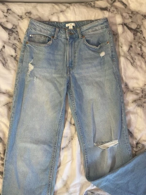 Ljusa mom jeans med slitningar - Säljer ett par ljusblå mom jeans med trendiga slitningar på låret och knät. Jeansen har hög midja, raka ben och klassiska fem fickor. Perfekta för en avslappnad och cool stil. Materialet är denim i 100% bomull.