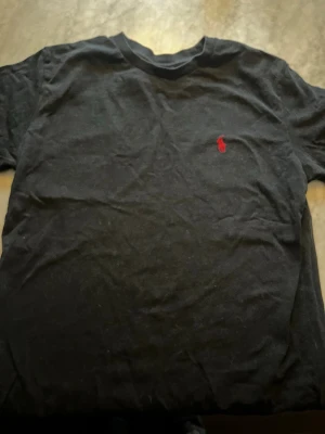 Svart t-shirt från Ralph Lauren - Svart t-shirt från Ralph Lauren med klassisk passform och korta ärmar. T-shirten har en liten röd broderad logga på bröstet och är tillverkad i mjuk bomull. Perfekt till jeans eller shorts för en enkel och stilren look.