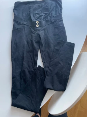 Svarta mammaleggings från Freddy WR.UP - Svarta leggings från Freddy WR.UP med hög midja och två dekorativa knappar framtill. Stretchigt material som formar sig efter kroppen och ger en snygg siluett. Perfekta för dig som vill ha både stil och komfort. Använda en gång som nya. 
